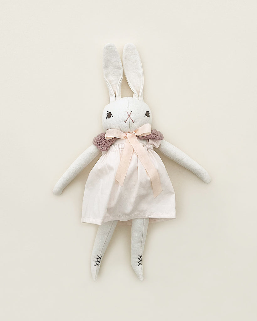 A Polka Dot Club Medium Cream Rabbit Hand Knit Collar with long ears and hand embroidered facial features, wearing a pink dress and a knit scarf, displayed against a light beige background.