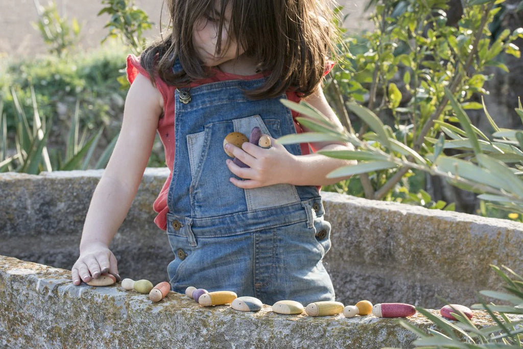 A young child in a denim overall plays with a row of colorful Grapat Insects on a concrete ledge outside, focusing intently on placing one among others.