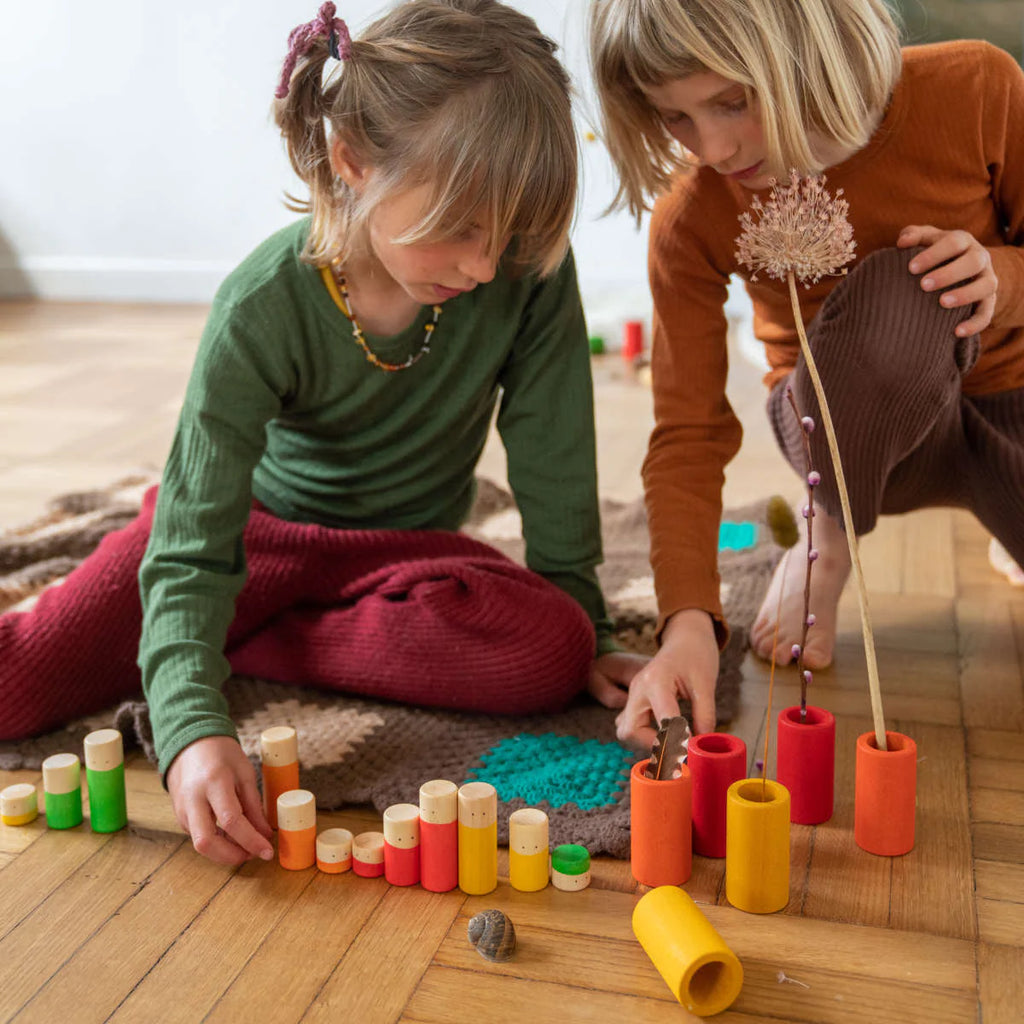 Two young children sit on a wooden floor, deeply focused as they play with the Grapat LA Non Basic Colors play set (36 pcs) and natural objects, engaging in creative, hands-on play that encourages divergent thinking.