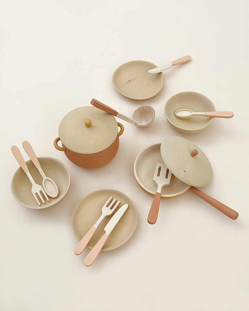 Handmade Wooden Kitchen Essentials - Flower featuring a large pot with handles and lid, three smaller pans with lids and long handles, two plates, a bowl, and various utensils including spoons, spatulas, forks, and knives—all laid out on a light surface. These handmade kitchen toys are crafted from non-toxic paint wooden toys.