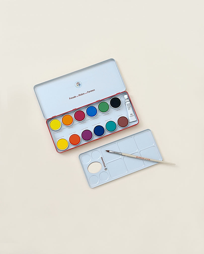 A set of Stockmar Opaque Colors - 12 Colors watercolor paints with 12 assorted colors, accompanied by two paint brushes, neatly placed on a beige background. The lid of the paint set displays the color mixing guide.