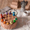 A chocolate frosted cake with colorful Grapat Together on top, surrounded by a coffee pot, empty glasses, and cardboard boxes, set on a textured beige cloth.