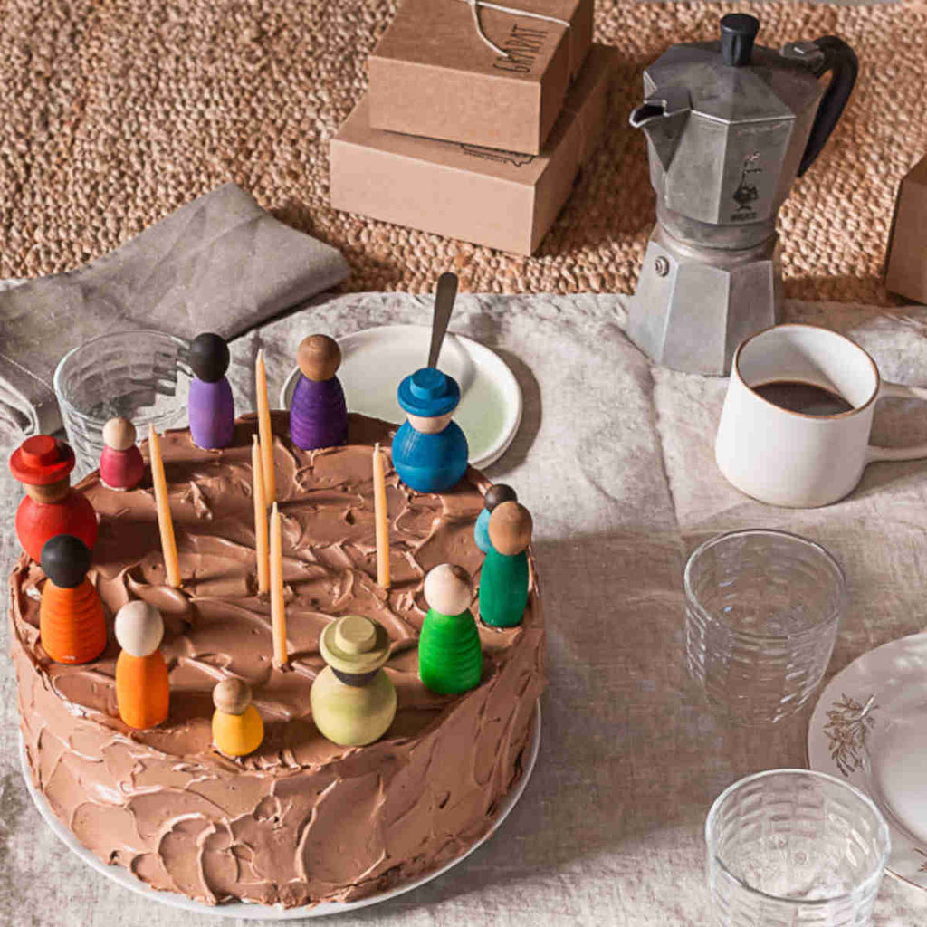 A chocolate frosted cake with colorful Grapat Together on top, surrounded by a coffee pot, empty glasses, and cardboard boxes, set on a textured beige cloth.