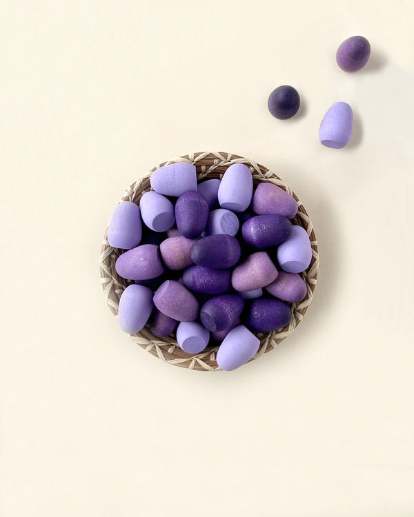 A variety of purple and lilac-colored stones arranged in a small woven basket on a light beige background, with a few Grapat Mandala Eggs scattered around the basket.