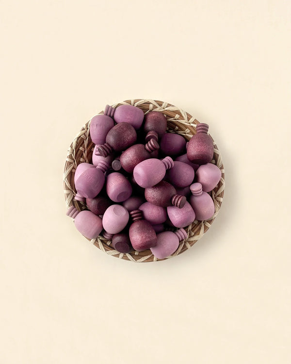 A wicker basket filled with various shaped and sized Grapat Mandala Pineapples, all painted in shades of purple, placed on a light beige background.
