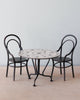 A Maileg Bistro Table With Chairs with a white, patterned top and black legs is flanked by two black, metal chairs with curved backs and no cushions. The setup sits on a light wood floor against a plain, pale blue background, perfect for an intimate Sunday supper.