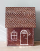 A small decorative house made from gingerbread with white icing details on the roof, a window, and a door. The roof is covered in scalloped icing patterns, and the door is marked with the number "24." This charming Maileg Cardboard Gingerbread House sits on a light wooden surface, perfect for holding holiday surprises.