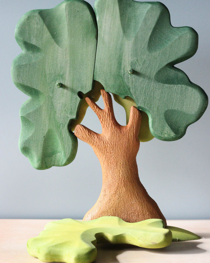 A handmade Extra Large Wooden Tree with a detailed trunk and large green leaves, standing on a table with a smaller green leaf piece in front. The background is plain and light-colored.