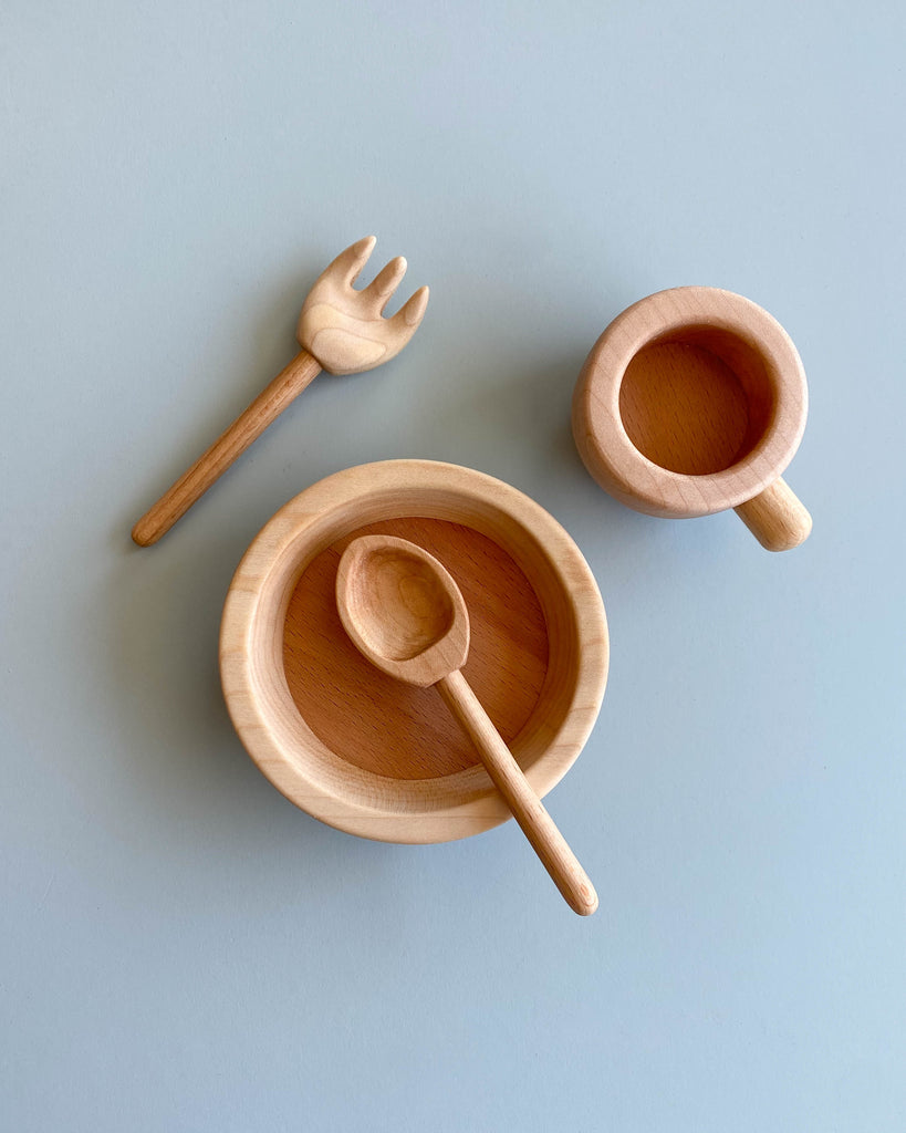 A flat lay of the Handmade Pretend Dinnerware Set with a natural wood finish on a pale blue background, featuring a bowl with a spoon, a cup, and a fork, all arranged neatly and symmetrically.