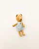 A small, worn Maileg Teddy Baby in a soft linen, blue and white checkered dress with a stitched heart on its chest, lying against a plain light background.