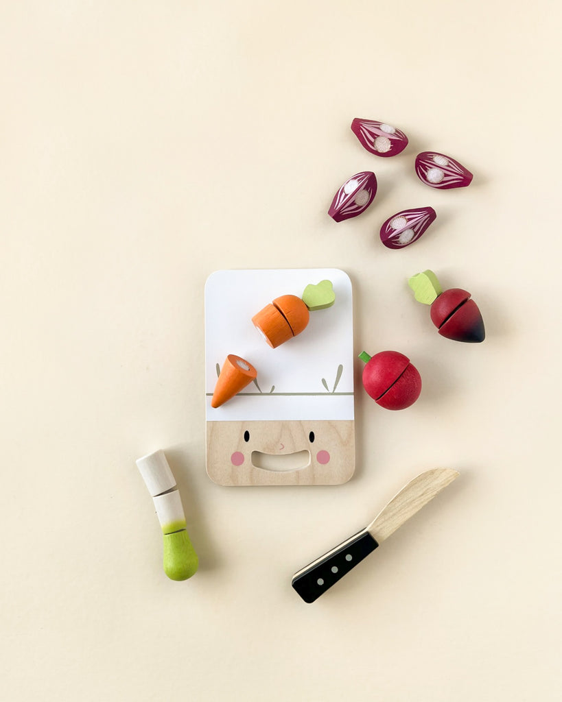 A playful flat lay of sustainable rubber wood toy vegetables arranged around a smiling Mini Chef Chopping Board, including carrots, radishes, and onions on a light background.