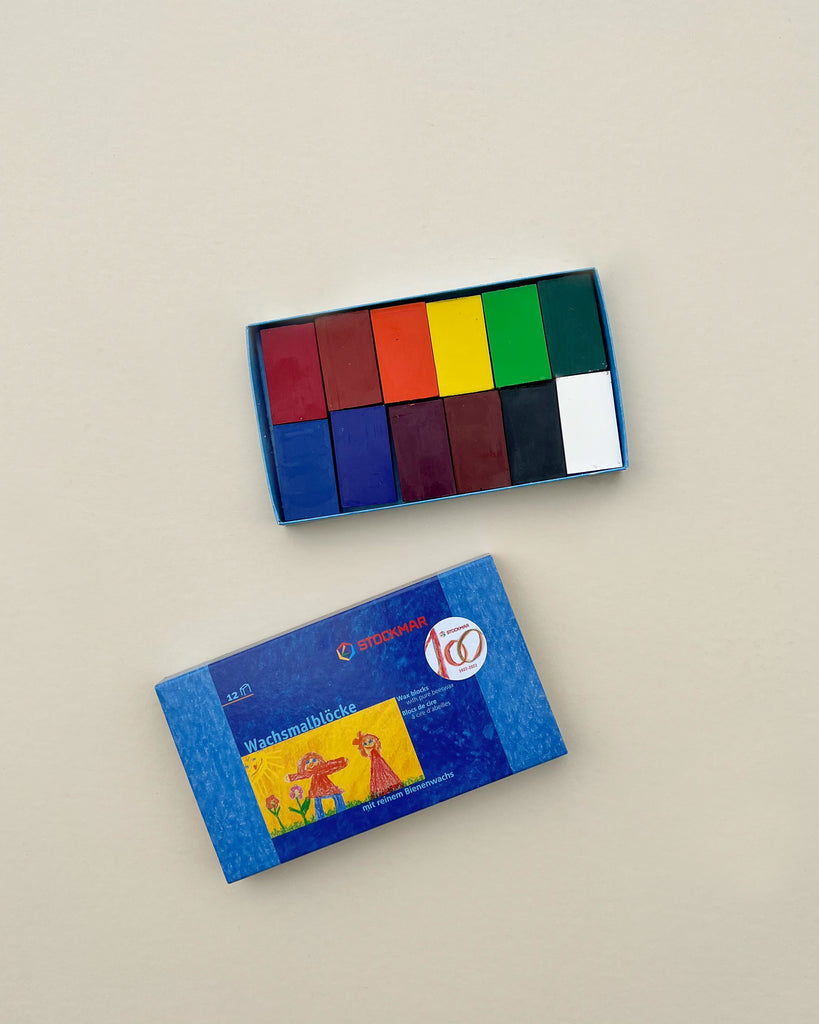 A set of colorful Stockmar Wax Block Crayons Box -12 Assorted in a blue cardboard box with a label depicting a child painting, placed on a plain beige background.