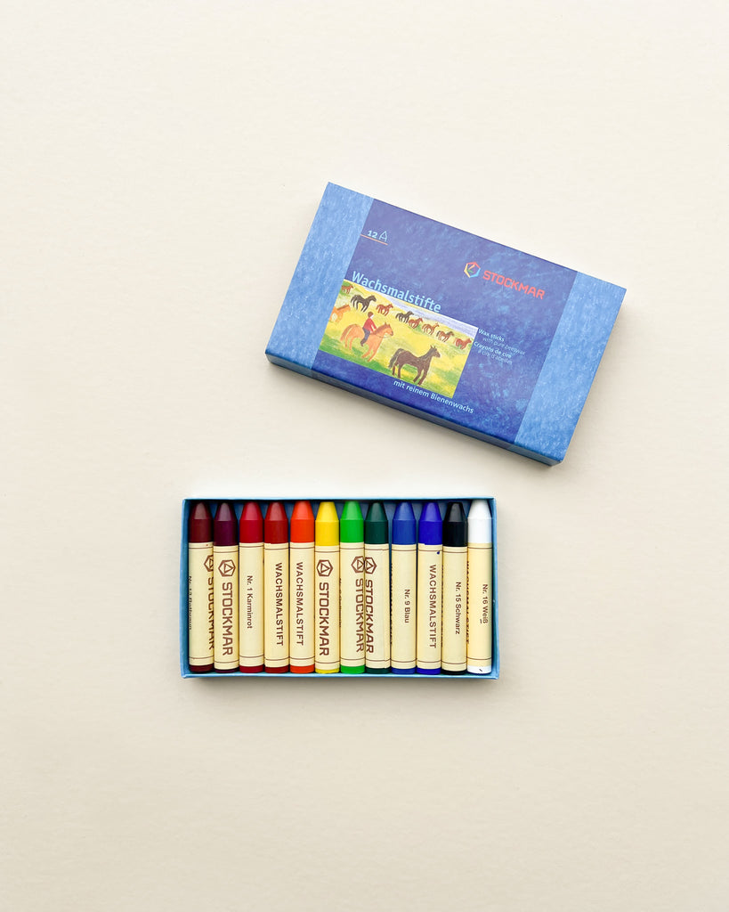 A box of 12 Stockmar Wax Stick Crayons lies open, displaying a range of vibrant colors neatly arranged next to the box cover, which features a colorful illustration of horses.