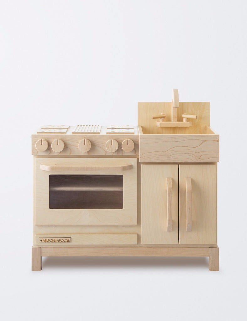 A Milton & Goose Wooden Play Kitchen featuring a stove, oven with a transparent door, and a sink with knobs, all Amish crafted in sustainable Baltic birch wood, against a plain white background.