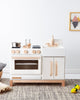 A Milton & Goose Wooden Play Kitchen - Made in USA, featuring a sustainable Baltic birch and white color scheme with a toy stove, oven, and sink, accessorized with pots, alongside a small wall hanging and plush