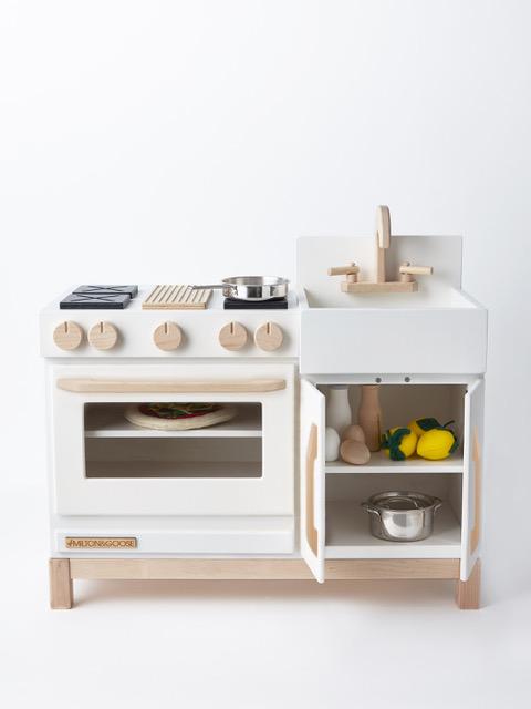 A small Milton & Goose Wooden Play Kitchen - Made in USA featuring an oven, stove, sink, and storage with toy fruits, vegetables, and utensils, set against a plain white background.