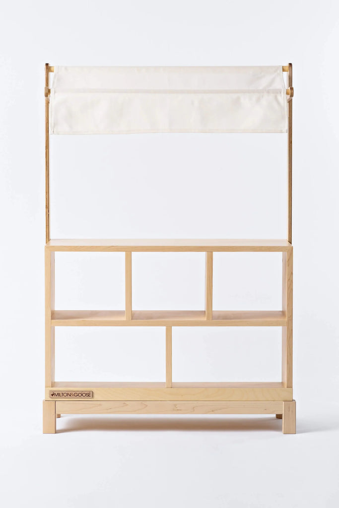 A Milton & Goose Market Stand with a plain canvas top and an open front displaying multiple shelves. This heirloom-quality design is handcrafted in the USA, featuring crisp lines and light-colored wood.