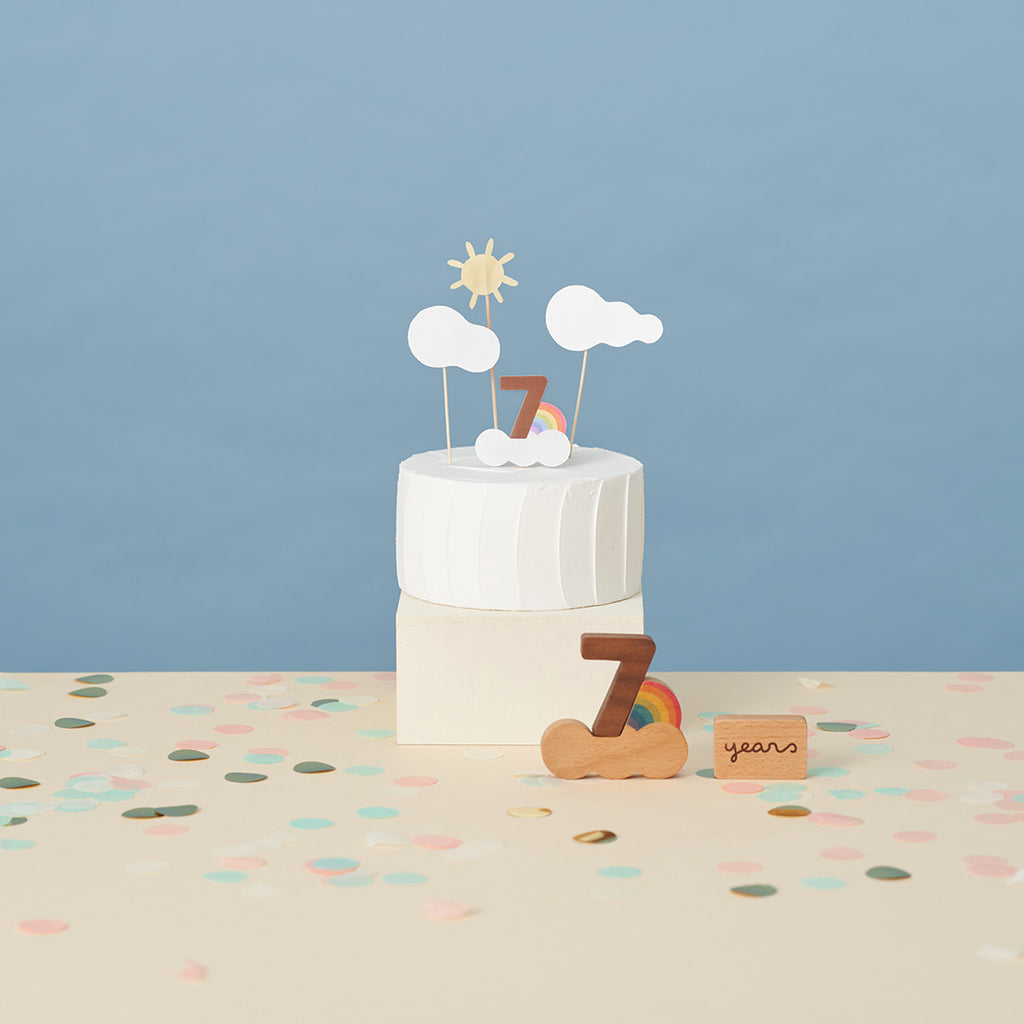 A minimalist and stylish 7th-anniversary celebration setup with a round white cake topped by decorative sun, clouds, and educational toys from Oioiooi Korea including the Numbers Play Block Set shaping the numeral.