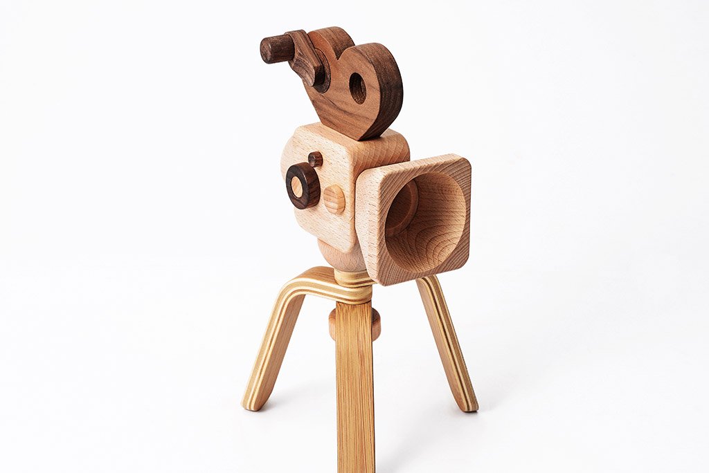 A Father’s Factory wooden toy camera with tripod legs, featuring detailed carvings to represent lenses and dials, set against a plain white background.