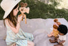 A young girl in a sunhat and blue dress engages in pretend-play with a Father’s Factory wooden toy camera with tripod, focusing it on two sitting dolls on a blanket under a tree's shade.