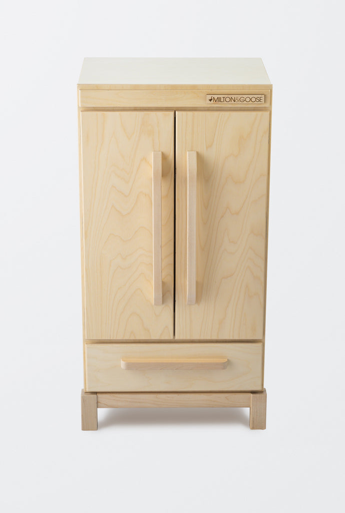 A Milton & Goose Pretend Refrigerator made in the USA, featuring two vertical handles on a pair of doors, with a single drawer beneath it. The furniture has a light natural wood finish and stands against a plain white wall.