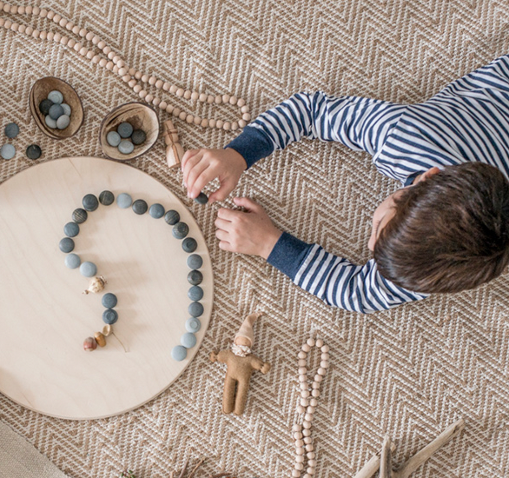 A child in a striped shirt sits on a textured rug, arranging Grapat Mandala Stones and beads into patterns on a circular wooden board made from wood sourced from sustainable forests, surrounded by crafting materials.