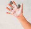 A child's hand against a light background, playfully tossing three small, Grapat Mandala Stones from sustainable forests in the air, capturing a moment of joyful movement.