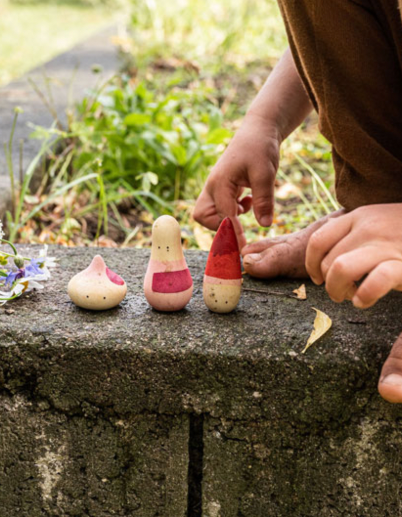 A person arranges three small, colorful Grapat Yay! Play Sets on a mossy stone surface outdoors, focusing on the one in the middle painted with red and white.