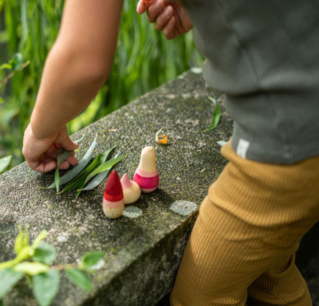A child in textured pants and a striped shirt engages in imaginative play with the small Grapat Yay! Play Set toys resembling gnome-like figures wearing pointy hats on a moss-covered stone outdoors, one figure adorned with a miniature sn