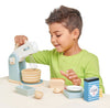 A young boy in a green shirt joyfully uses a Wooden Baking Set to pretend making a cake, with wooden bowls, eggs, and a flour box nearby, isolated on a white background.