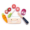 A smiling Mini Chef Chopping Board with a face, surrounded by colorful wooden play food items including a carrot, tomato, onion, and leek, and a wooden knife.