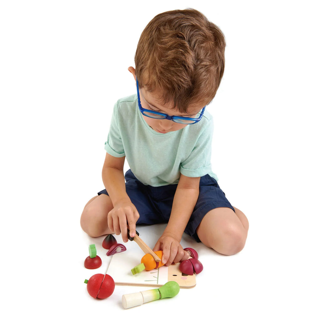 A young boy wearing blue glasses and casual clothing plays with a Mini Chef Chopping Board on a white background.