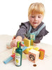 A young child playing with a Grocery Set, including boxes and cans, on a white table. The child is focused on a toy olive oil bottle.