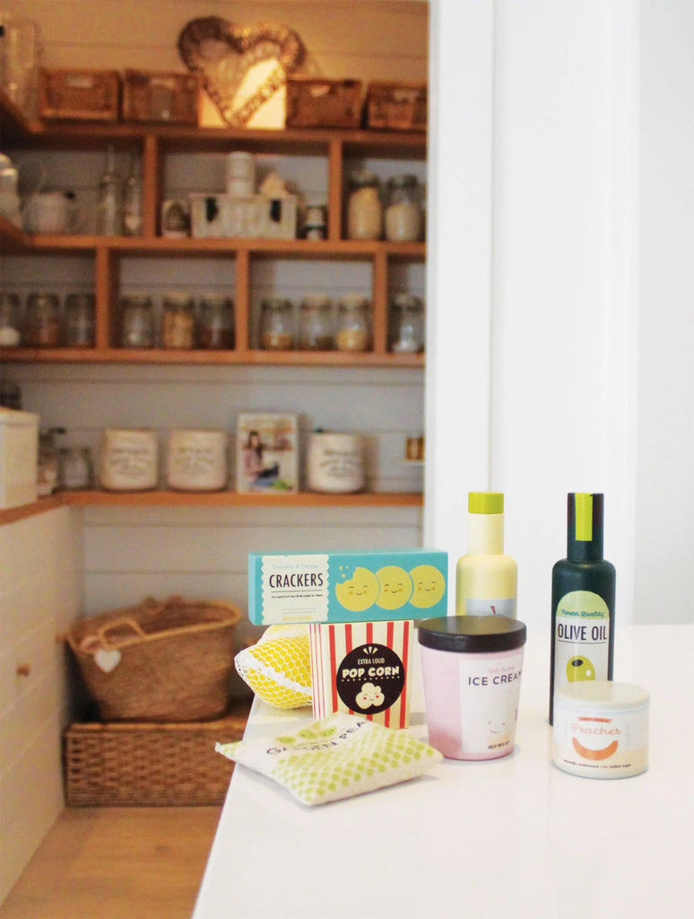 Various grocery set items like crackers, popcorn, and oils displayed on a kitchen countertop with open wooden shelves filled with baskets, toddler kitchen accessories, and kitchen essentials in the background.