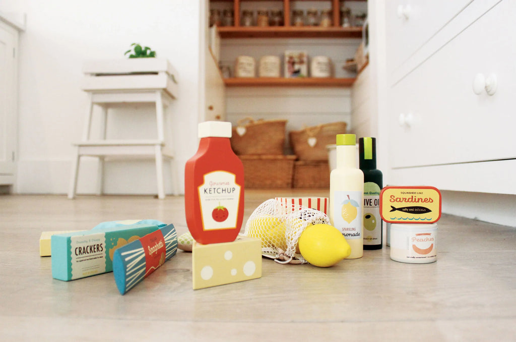 Various Grocery Set items like ketchup, vegetables, cheese, crackers, and sardines scattered on a kitchen floor with a white cupboard in the background.