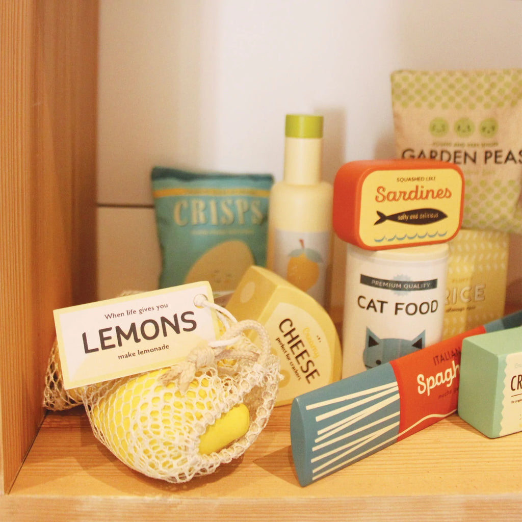 A variety of colorful, playfully packaged Grocery Set items, including fake cheese, lemons, spaghetti, and sardines, neatly arranged on a wooden shelf as part of a toddler kitchen accessories.