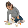 A young girl with a ponytail, wearing a striped top and denim skirt, engages in imaginative play with Stacking Garden Friends and a toy tree on a white background.
