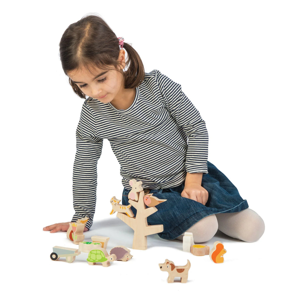 A young girl with a ponytail, wearing a striped top and denim skirt, engages in imaginative play with Stacking Garden Friends and a toy tree on a white background.