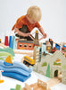 A young boy with blonde hair plays intently with a Mountain View Train set that includes buildings, vehicles, and landscapes, all spread out on the floor.