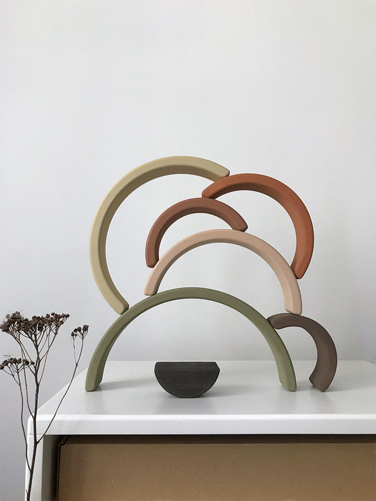 A minimalist sculpture featuring the Handmade Rainbow Stacker - Olive, with arc-shaped pieces stacked in a semi-circle arrangement on a white surface. The arcs, resembling a sophisticated stacking toy, come in various colors including green, beige, brown, and orange. Dried flowers are placed on the left side of the surface.