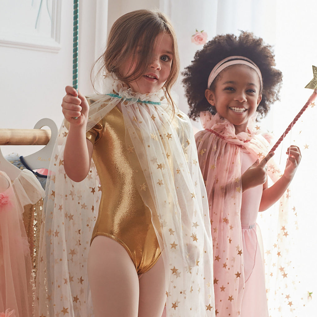 Two young girls playing dress-up, one dons a gold bodysuit and a Meri Meri White Tulle Star Cape Costume; the other is in a pink bodysuit and cape. Both are smiling, holding fairy w