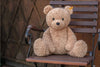 A small, fluffy, tan Steiff XL Jimmy teddy bear sitting on a wooden bench with a stone wall in the background.