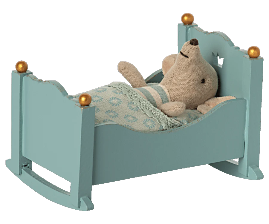 A plush teddy bear lying cozily under a blanket in a Maileg Miniature Cradle with golden knobs.