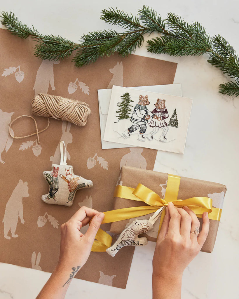 Hands tying a golden ribbon around a wrapped gift, including illustrated cards and holiday decorations, such as fir sprigs and the Coral & Tusk Ice Skater Bunny and Fox Ornament.