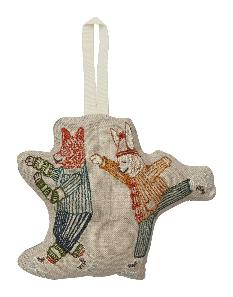 A Coral & Tusk Ice Skater Bunny and Fox Ornament depicting two anthropomorphic cats in figure skating outfits, ice skating, with one performing a jump and the other a spin, crafted in detailed stitching on a beige background.
