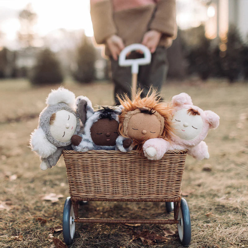A child pushes a small wicker pram filled with four Olli Ella Zebra Mini Dolls—a sheep, a collectible doll with brown skin, a fairy, and a cat—on a grassy area with trees.