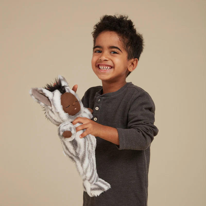 A joyful young boy with curly hair, wearing a gray shirt, holds and shows a Olli Ella Zebra Mini Doll with a smile against a neutral background.
