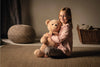 A young girl with a joyful smile, sitting cross-legged on the floor, happily embraces a large Steiff XL Jimmy Teddy Bear, 22 Inches with the signature Button in Ear in a warmly lit room.