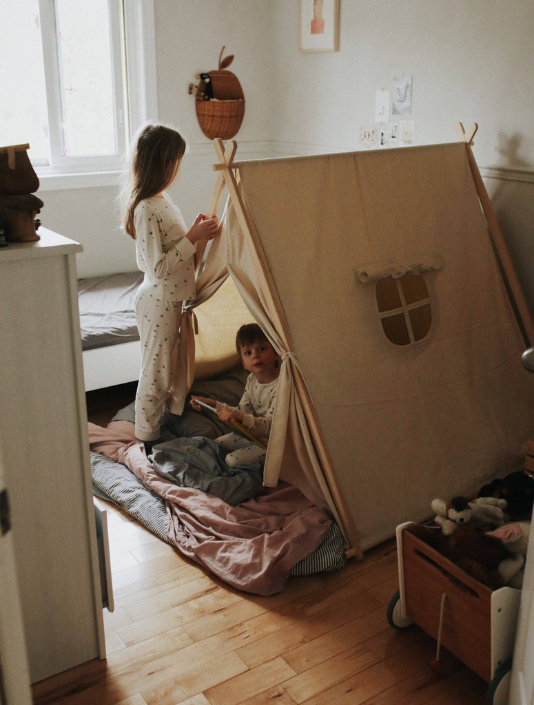 Two children play inside an Indoor/Outdoor Play Tent tent in a cozy, warmly lit bedroom, surrounded by toys and soft pillows, creating a playful and inviting atmosphere.