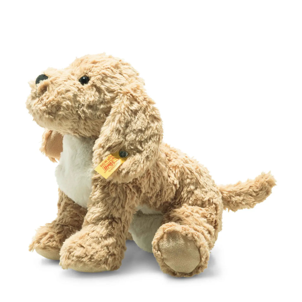 A Steiff, Jimmy Berno Goldendoodle Puppy, 10" plush toy with a tan, curly fur texture, sitting in a playful pose, isolated on a white background. The toy features black eyes and a small, distinguishable